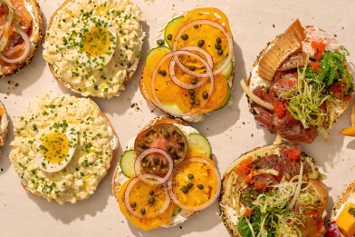 Overhead view of several open-face bagels topped with different spreads, including egg salad with half a boiled egg, heirloom tomatoes with cucumbers and capers, and a poke-style mix with seaweed salad, smoked fish, onions, and roe, all arranged on parchment.