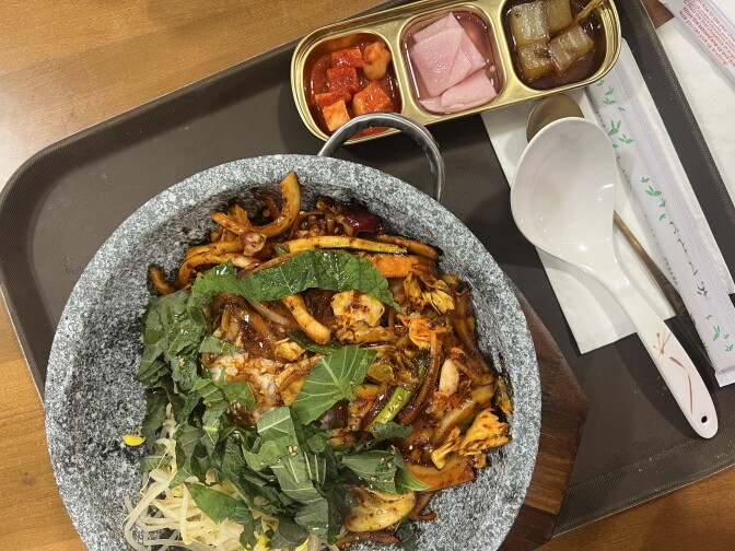 A grey pot on a brown tray alongside pickles in a small gold tray. The grey pot is stone and contains caramel-colored squid and fried onions alongside green perilla leaves and yellow bean sprouts.  
