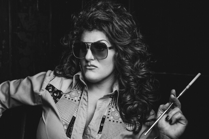 Black and white image of a light-skinned woman wearing sunglasses and holding a cigarette in a holder. 