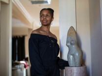 A Black woman wearing an off-the-shoulder black blouse looks off to the left of the frame. She stands next to a sculpture of a woman's torso.