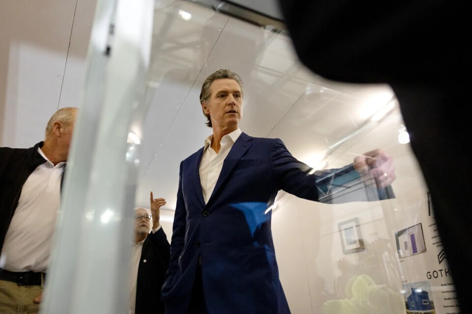 A man who appears to be white and in his 40s or 50s points to something on a glass door. He wears a navy suit and white shirt open at the collar. His hair is gray and slicked back. Two other men can be seen behind him, who appear to be white and in their 60s. He is indoors. 