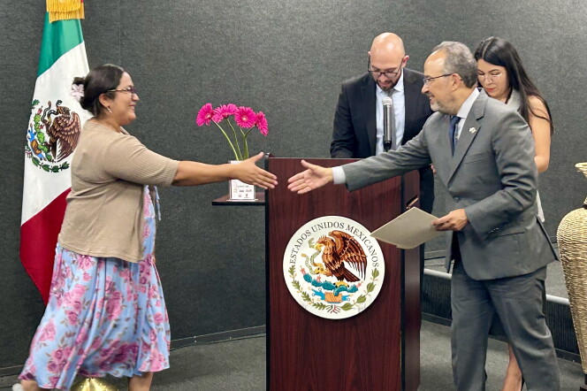 A male presenting person and a female presenting person reach to shake each other's hands. The flag of Mexico is behind the woman.