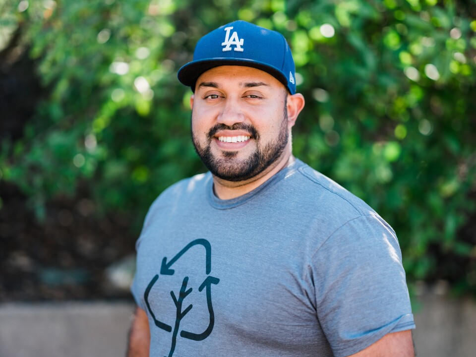 A headshot of a man with light brown skin wearing a blue LA dodgers hat and grey shirt with a recycling motif on the front. THe man smiles and has a black short beard and mustache. The background is blurred greenery.  