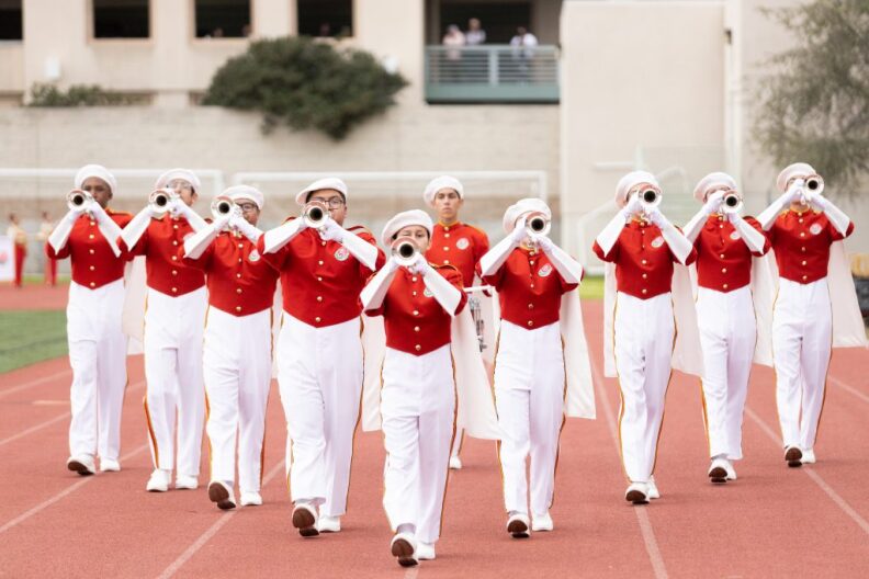 Trumpeteers wearing white and red uniforms march in formation on a track.