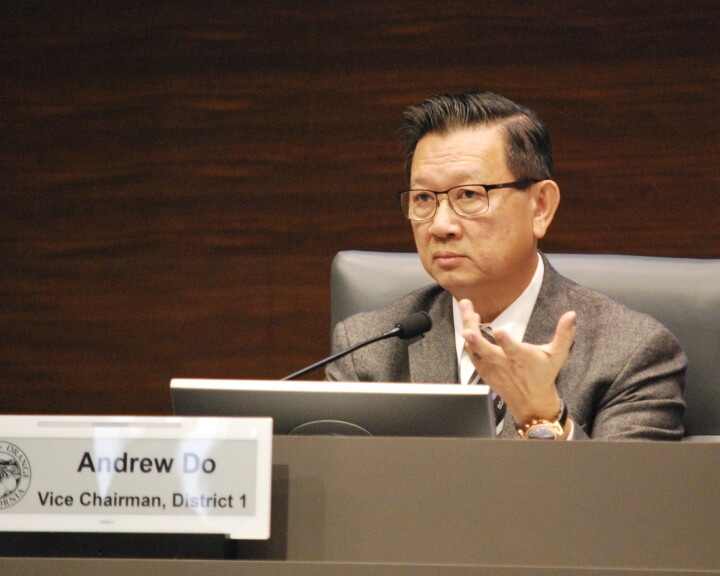 A man in a chair wearing a suit jacket, tie and glasses looks forward with a microphone in front of him. A sign in front has the official seal of the County of Orange and states "Andrew Do, Vice Chairman, District 1."