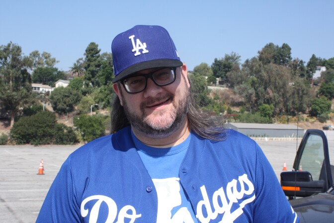 A man wearing a Dodgers hat and Dodgers short is smiling into the camera. He's got brown hair and glasses.