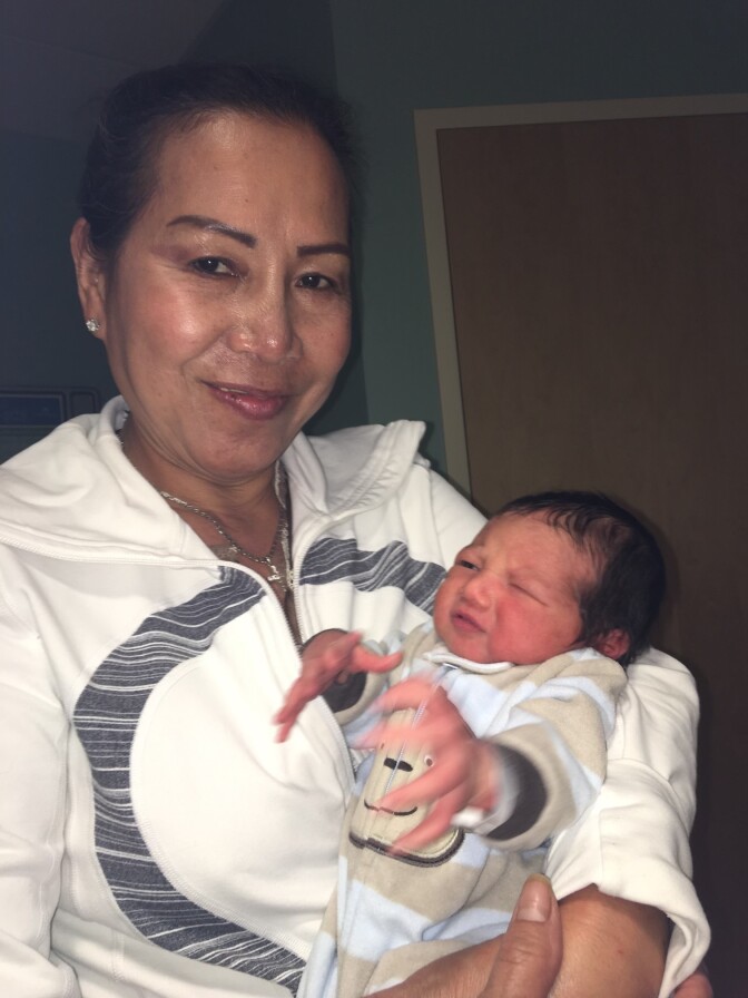 A Vietnamese American woman in her late 50s wearing a white jacket holds a newborn baby. 