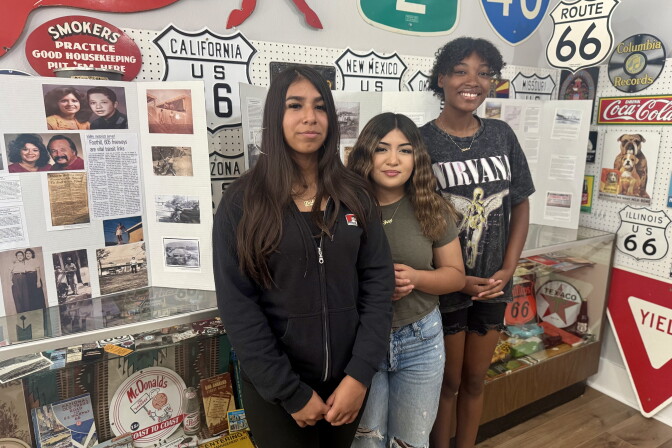 Three girls with varying skin tones stand in front of a series of tri-fold posters that are covered in photos and old news clippings.