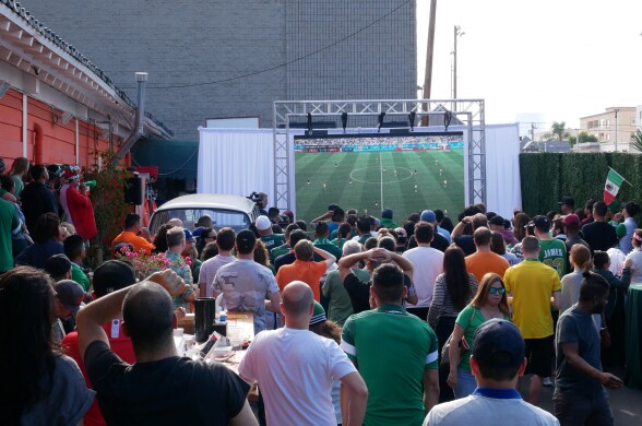 A group of people standing outside, watching a large scree with a soccer game playing on it.