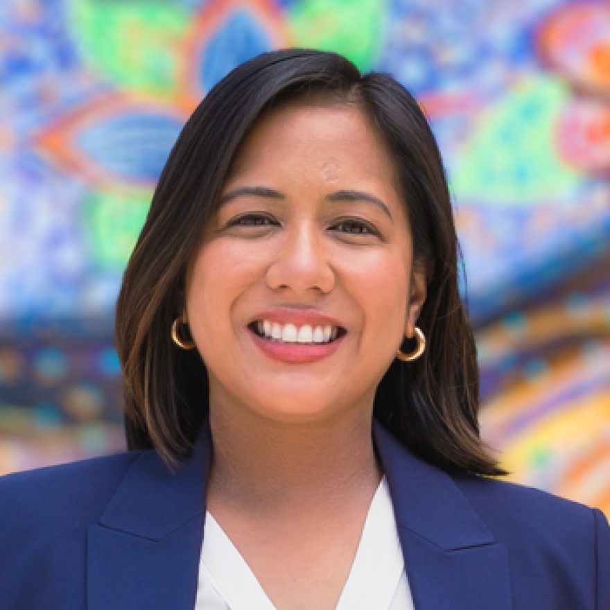 A woman with shoulder-length dark brown hair, wearing hoop earrings and a blue blazer smiles at the camera. She's standing against a colorful backdrop of painted flowers
