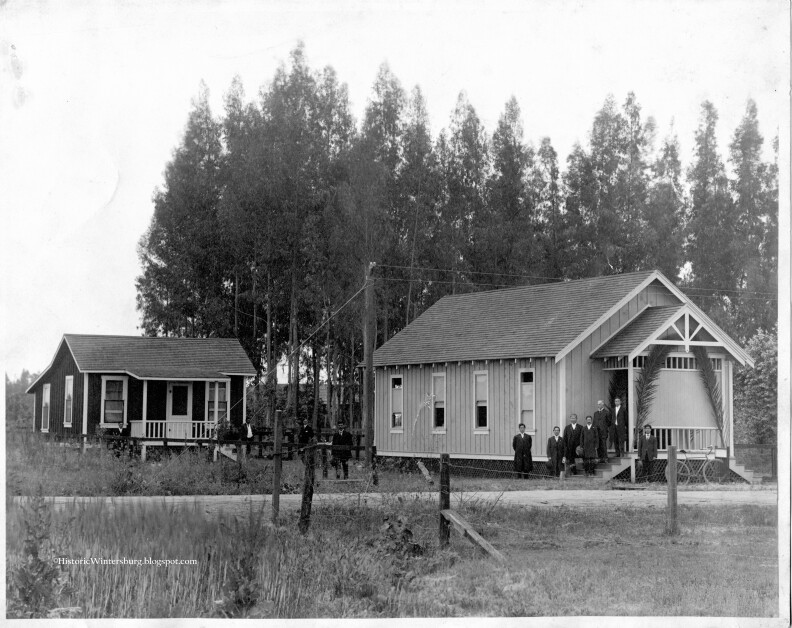 A black and white photo of two small wooden structures, a manse and a mission. Asian men in suits stand outside the larger mission.
