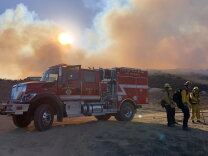 A firefighting truck with personnel standing next to it. Smoke clouds are seen in the background.