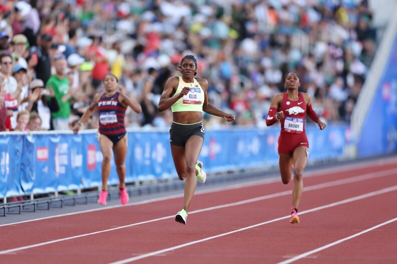 Three woman with dark skin run alongside each other on a track, wearing different colored uniforms. A crowd can be seen in the background cheering.