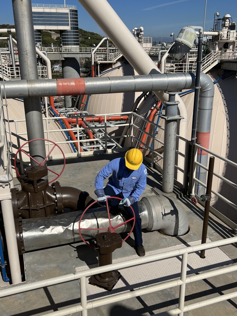 A man wearing a blue shirt and yellow hard hat turns a wheel in on top of an industrial facility.