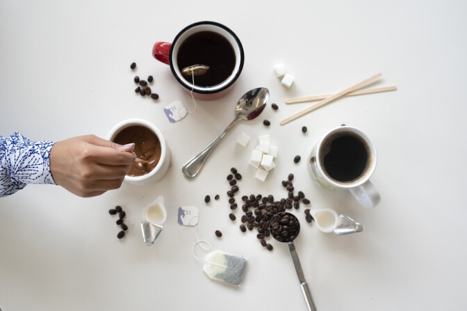 A hand stirs a coffee on a white table that also shows tea and coffee beans