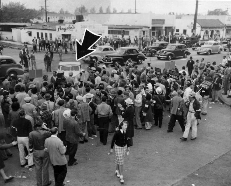 A crowd is gathered in the street in front of a light-colored car. A restaurant, Angelo's Cafe, can be seen across the street. Several other buildings and trees can be seen in the background. Picket signs read "End lockout says U.S. Govt. to producers."