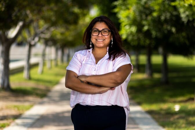 A woman dressed in a short-sleeved striped top and black pants stands smiling with her arms folded on a tree-lined walkway during the day, at Pasadena City College.