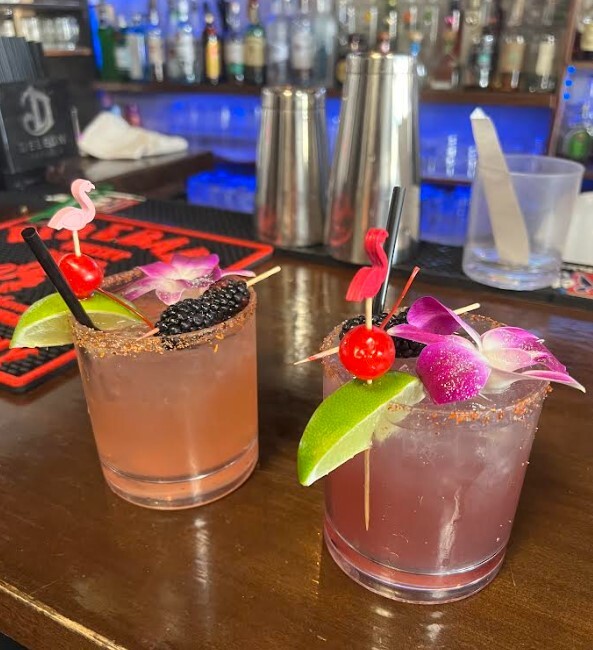 Two cocktails — both accented with a cherry-and-lime flamingo skewer — are positioned next to each other on a brown wooden bar top.