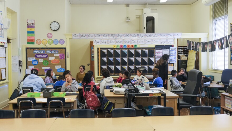A classroom at Carson Street Elementary. There are 15 visible third grade students sitting at desks. The walls are a cream color. There is a corkboard with letters that spell out "Mindset Matters" and depictions of cursive letters lining the wall.