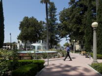 A man wearing black pants and a blue long sleeve walks across a plaza with a large water fountain at its center. 