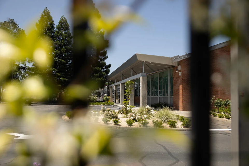A brick building with a sloped roof and concrete canopy over a walkway, with a parking lot outside, photographed through a fence. The parking lot is partially obscured by an out-of-focus shrub and part of the fence.