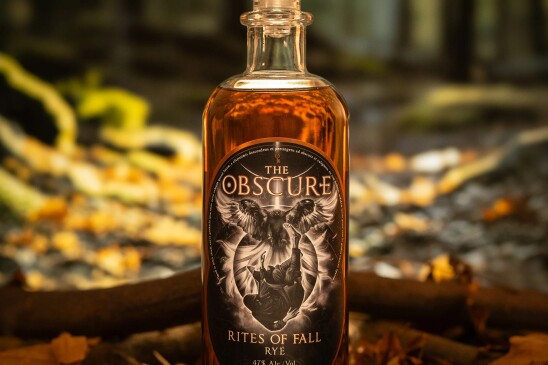 A bottle of Rites of Fall rye whiskey by The Obscure is displayed among fall leaves and a fallen tree branch.