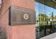 The exterior of a building has a terra cotta-colored stone wall with the words : County of Orange County Administration South on a plaque dated 2029. The names of county officials are on the sign. A glass wall reflects U.S. California and the county flag.