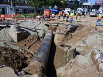 A section of a city street has been dug up, creating a deep trench with an exposed pipe running down the middle. 