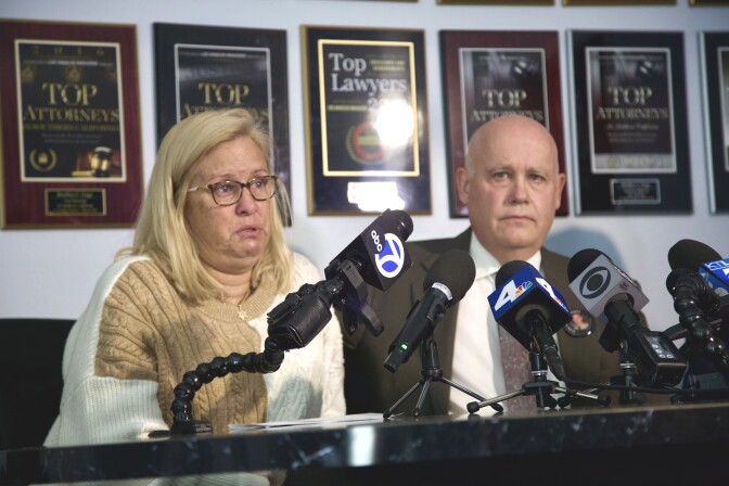A blonde woman wearing glasses is visibly emotional as she sits in front of a table with several news microphones. A man in a suit is sitting next to her.