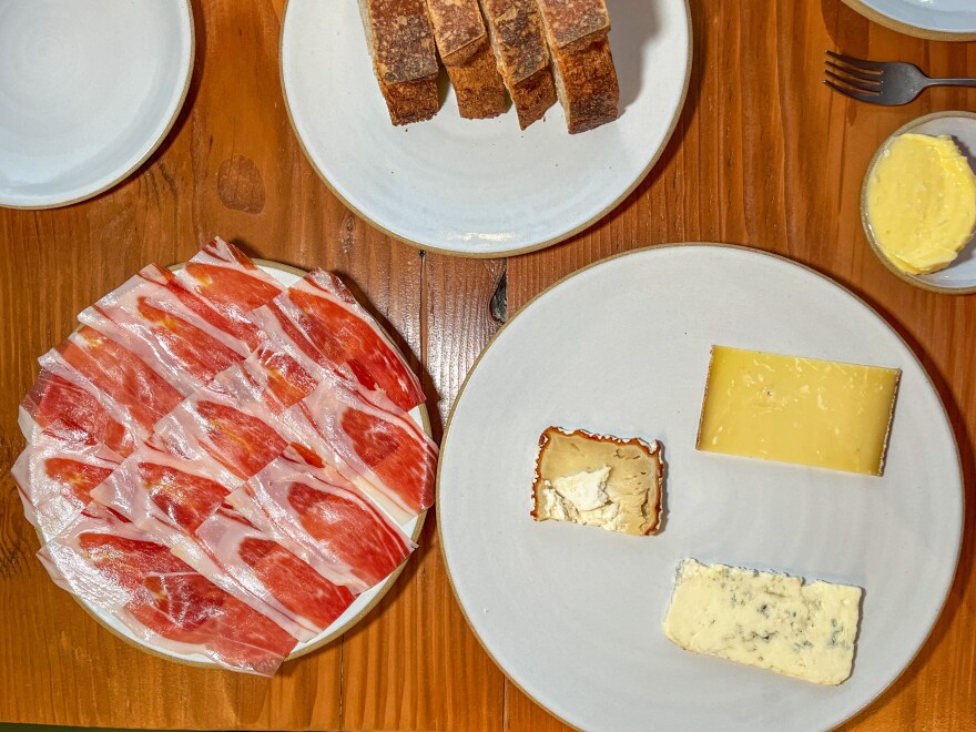 A series of white ceramic plates are arranged on a medium-dark wooden tabletop. One has overlapping slices of red and white cured ham, another has three chunks of different kinds of cheeses; and the last has four slices of crusty brown sourdough bread
