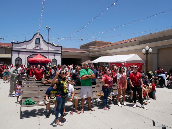 A group of people stand in a courtyard. A women wearing a blue shirt and sunglasses has her hands up and is holding