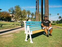 A male presenting person sits on a bench. The bench is painted in bright blue and yellow.