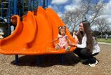 A toddler girl with pigtails sits at the bottom of a slide at a park next to her mother.