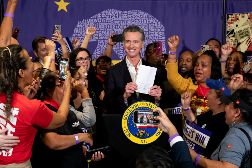 A large crowd of people, many with their fists raised, gather around Governor Gavin Newsom, who is holding a piece of paper and standing in front of a podium with a round logo that reads Seal of the Governor of the State of California