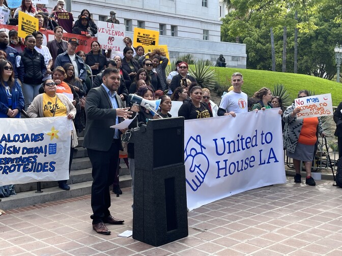A man with medium-tone skin wears a suit with no tie as he stands at a lectern in front of group of people, some holding a "United to House LA" banner