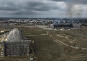 In the foreground, a massive hangar sits intact. To the right of it, a twin structure is in ruins, with smoke still rising from the hull. All around it is wide open space to one side, and signs of suburban development to the other.