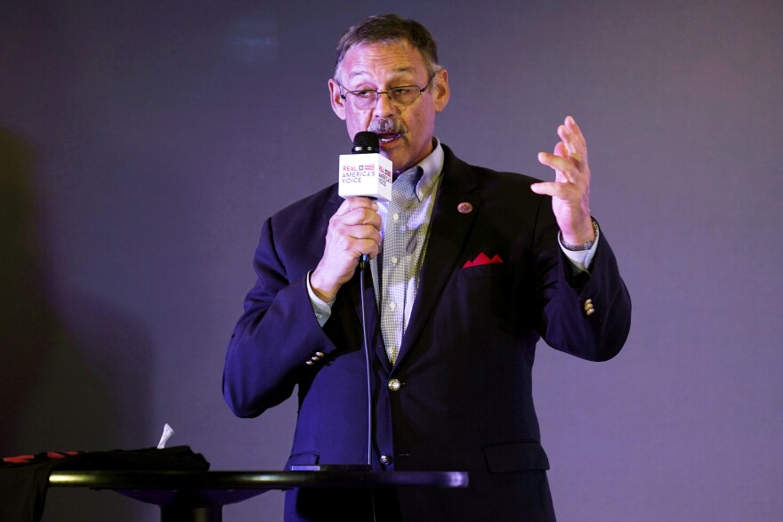 A man with a narrow moustache and dark suit, with a red pocket square and no tie, holds a microphone to speak.