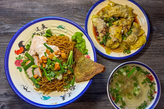 An overhead view of the full order at Mai Phát Mì Gia — a large rooster-print plate of dry egg noodles topped with char siu pork, crispy lard, green onion, and a fried wonton, alongside a bowl of plump sủi cảo dumplings in soy sauce and a small clear broth soup.