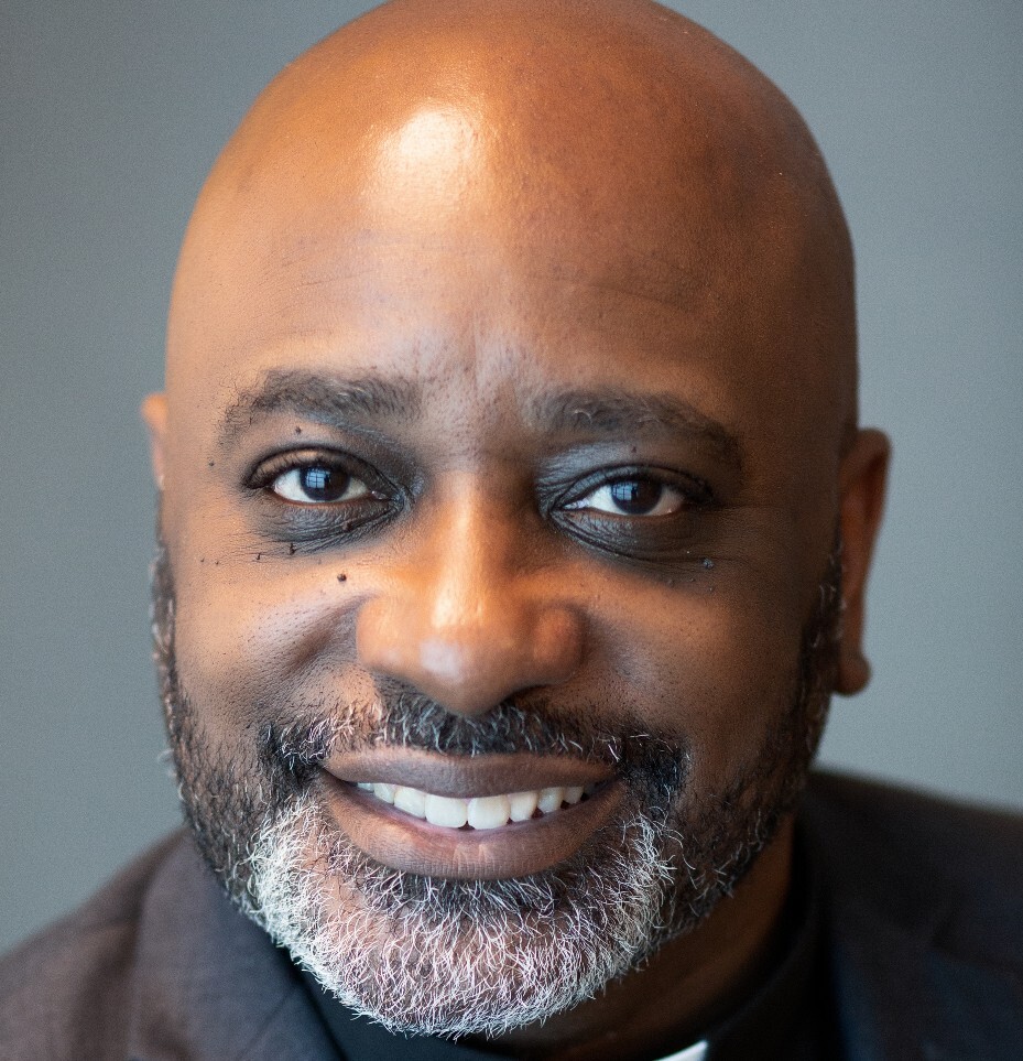 The Rev. Dr. James Thomas, a Black man with dark eyes, a close-cropped beard and moustache with grey in the beard below his mouth, smiles at the camera. He is bald.