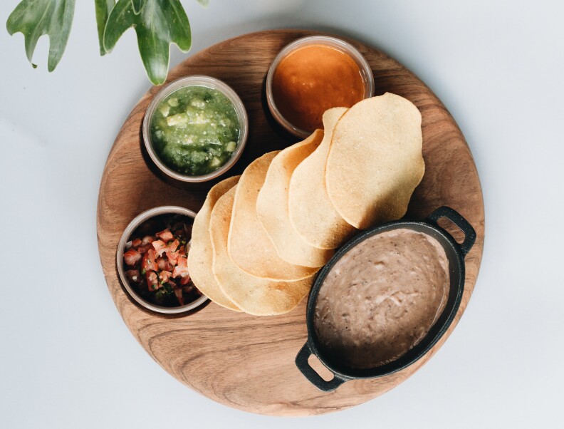 a wooden circular plate with torillas in the middle, surrounded by guacamole, salsas and refried beans.