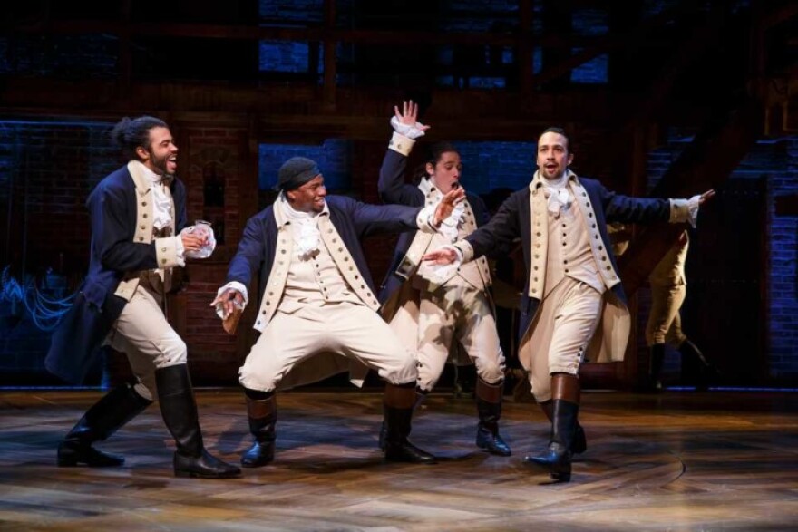 4 men in old timey, American Revolution uniforms, dance and sing on a stage