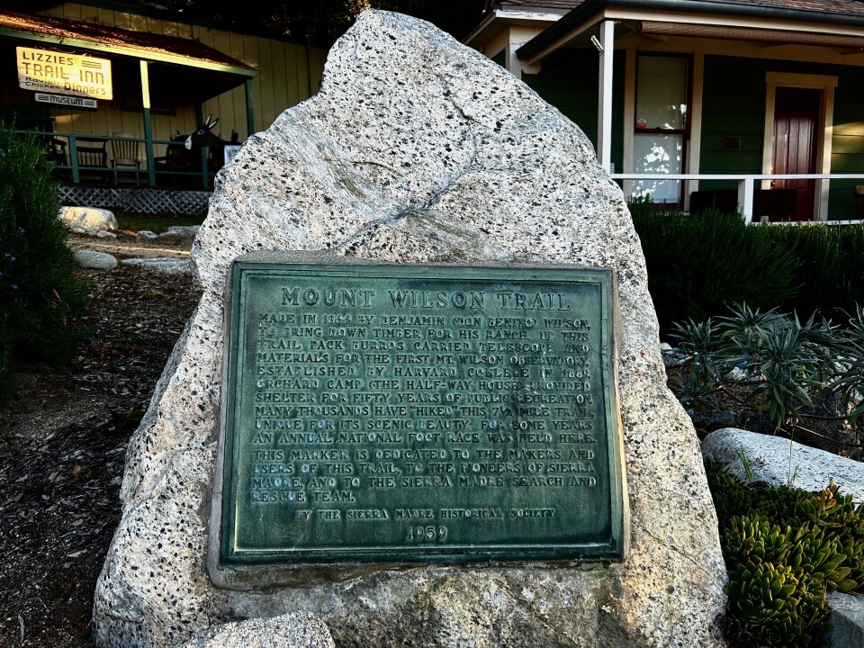 A wide image of a large boulder with a turquoise, rusted plaque that reads "Mount Wilson Trail." Small wood buildings and a museum are behind. 