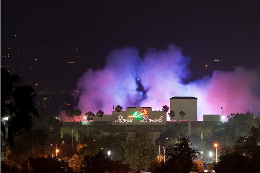 Clouds of purple-tinged smoke rise from the Rose Bowl with the stadium's iconic neon sign featuring a red rose visible in the foreground.