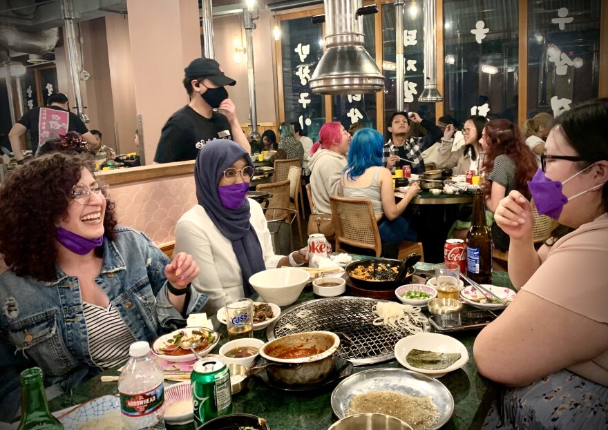 Three young women in their 20s laugh as they dine at a crowded restaurant with a Korean BBQ grill at their table, surrounded by plates of food.