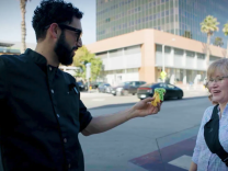 A man with a light skin tone and dark hair, wearing glasses and a black chef's coat, extends a colorful taco toward a woman with light hair and a light skin tone on a sunlit Los Angeles sidewalk. The woman, wearing a plaid shirt with a crossbody bag, smiles mid-conversation as office buildings and palm trees line the street behind them.