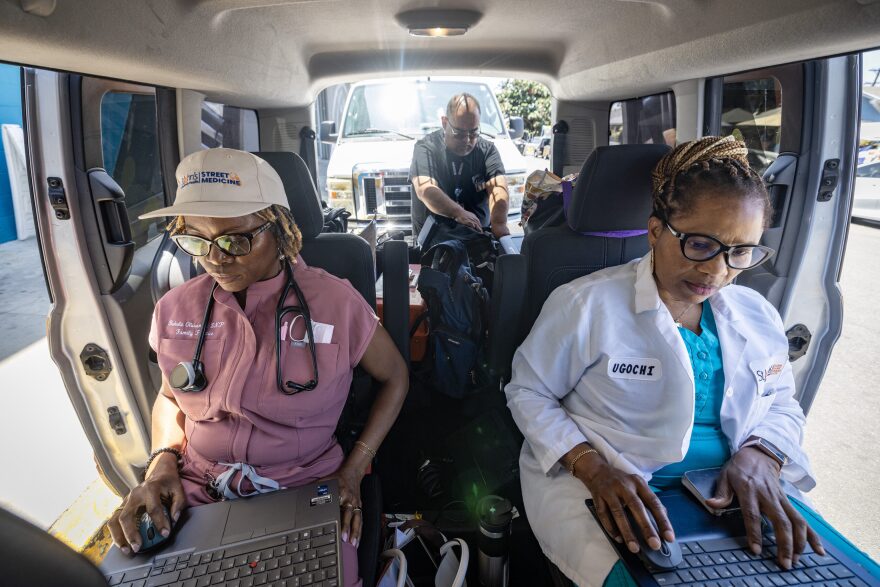Two women sit in the backseat of a van while working on laptops. Women on the left wears a white baseball cap, mauve scrubs and has a stethoscope around her neck. Woman on the right wears a white medical lab coat over a turquoise top.