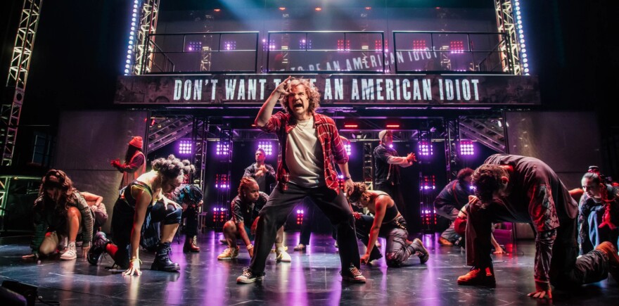 The full 'American Idiot' ensemble performs the titular song on stage. The lead singer stands center stage in a flannel shirt, white tee, and slacks as he sings. Behind him the lyrics "don't want to be an American idiot" are displayed across a steel awning.