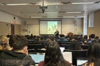Rows of students with laptops face a light-skinned woman with short hair in front of a projection screen. The screen features white text on a black background (illegible), along with Pablo Picasso's 1955 sketch of Don Quixote and and his sidekick, Sancho Panza.