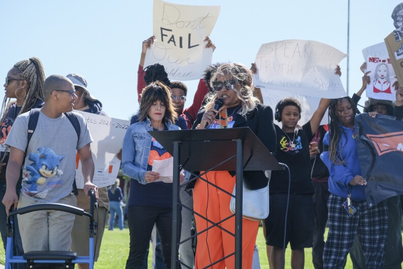 Teenagers with dark skin tone stand around a woman with dark skin tone speaking at a podium set up on the lawn of a high school campus.