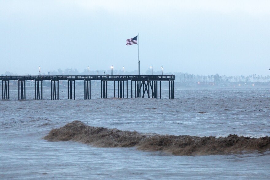 A pier on the left of the frame sticks out towards the middle of frame. Towards the end of the pier an American flag blows in the wind. The ocean water is gray and brown from the stormy day.